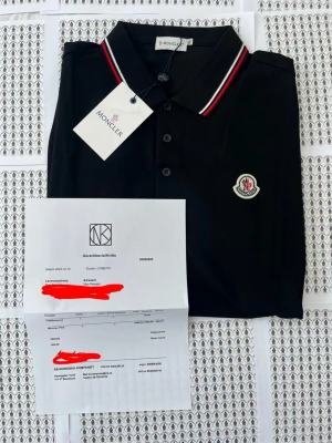 Svart Moncler piké med rödvit krage - Snygg svart pikétröja från Moncler med klassisk krage och knappar framtill. Kragen har röda och vita ränder för en sportig touch och bröstet pryds av Monclers ikoniska logga. Perfekt för dig som gillar stilrena märkeskläder.