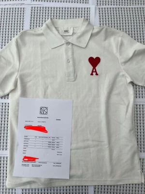 Vit pikétröja från Ami med rött hjärta - Vit pikétröja från Ami med klassisk krage och knäppning framtill. På bröstet finns ett stort rött broderat hjärta med bokstaven A. Tröjan är gjord i bomull och har en clean, stilren look som passar till mycket.