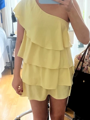 Gul one shoulder volangklänning - Säljer en ljusgul kortklänning med flera lager volanger och oneshoulder-design. Klänningen har en luftig och flowy passform, perfekt för sommarens alla tillfällen. Materialet känns lätt och svalt mot huden.