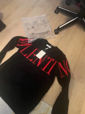 Svart stickad tröja från Valentino - Svart stickad tröja från Valentino med stora röda bokstäver som bildar loggan över bröstet och armarna. Tröjan har rund halsringning och långa ärmar. Perfekt för dig som gillar exklusiva och trendiga plagg med statement-tryck.