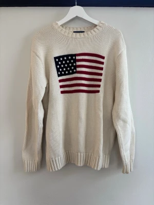 Vit stickad tröja med USA-flagga - Vit stickad tröja med rund hals och ribbade muddar. På bröstet finns en stor amerikansk flagga i rött, vitt och blått. Tröjan har en klassisk passform och är gjord i ett tjockare garn som känns både mjukt och rejält.