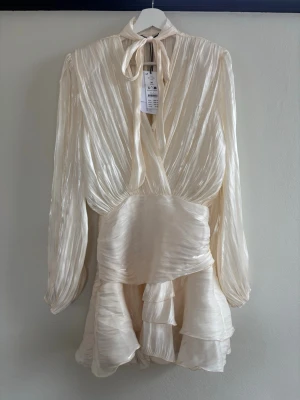 Krämvit omlottklänning med volanger - Säljer en elegant krämvit kortklänning i glansigt material med omlottdesign, långa ärmar och volangdetaljer. Klänningen har knytband i halsen och en lätt plisserad struktur som ger en lyxig känsla. Perfekt för dig som vill sticka ut med en chic och trendig look.