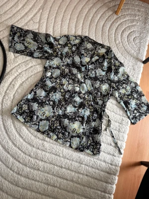 Blommig omlottblus i mesh - Snygg svart meshblus med blommigt mönster i blått, gult och vitt. Omlottmodell med knytband i sidan och trekvartsärm. Perfekt för dig som gillar transparenta material och trendiga prints.