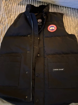 Svart dunväst från Canada Goose - Säljer en svart dunväst från Canada Goose med klassisk logga på bröstet. Västen har hög krage, två stora fickor framtill och dragkedja med tryckknappar. Insidan har en meshficka. Perfekt för lager på lager och riktigt snygg streetstil.