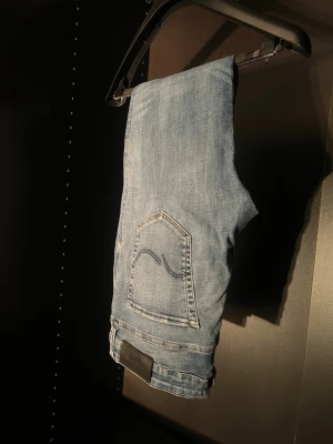 Ljusblå raka jeans från Jack & Jones - Säljer ett par ljusblå jeans från Jack & Jones med klassisk femficksdesign och raka ben. Jeansen har en snygg tvättad look och subtila slitningar för en avslappnad vibe. Tillverkade i bomull med Nudies ikoniska bakficka. Perfekta till vardagsstilen!
