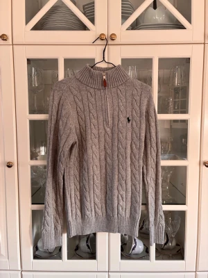 Grå kabelstickad tröja Polo Ralph Lauren - Snygg grå kabelstickad tröja från Polo Ralph Lauren med halv dragkedja och hög krage. Tröjan har det klassiska broderade logotypen på bröstet och ribbade muddar vid ärmslut och nederkant. Perfekt för lager-på-lager och stilren look.