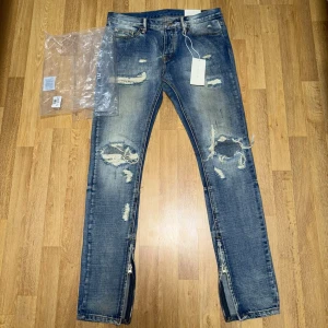 Säljer ett par MNML Repaired Jeans Blue – Size W32 - Jeansen är helt nya och endast testade. Inga skador eller slitage.  Trendiga jeans med ripped/repaired design och dragkedjor vid anklarna som ger en modern streetwear-look. Original tags medföljer.  Size: W32 🏷️ Rabatt vid köp av flera plagg!