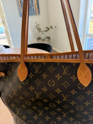 Louis Vuitton Neverfull Monogram - Säljer en klassisk Louis Vuitton Neverfull handväska i brunt monogramcanvas med ljusbruna läderdetaljer och långa axelband. Insidan är randig i beige och svart med guldfärgade metalldetaljer och en innerficka med dragkedja. 