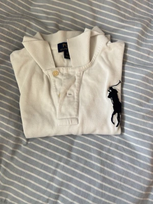 Vit pikétröja från Polo Ralph Lauren - Klassisk vit pikétröja från Polo Ralph Lauren med svart broderad logga på bröstet och nummerdetalj på ärmen. Tröjan har krage, knappar framtill och är tillverkad i bomull. Perfekt för en stilren och sportig look.