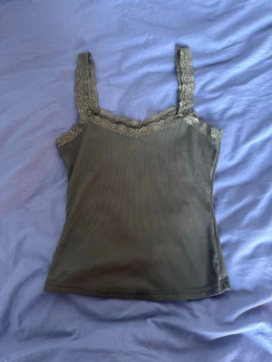 Olivgrön ribbad linnetopp med spets - Snygg olivgrön ribbad linnetopp med breda axelband och dekorativ spets längs ringningen och banden. Perfekt för lager på lager eller till jeans. Mjuk och stretchig känsla.