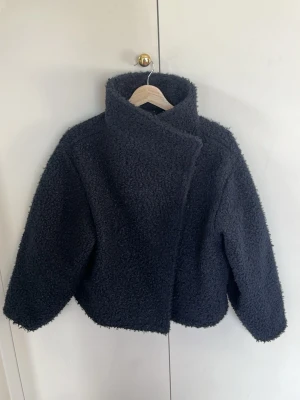 Marinblå kappa - Mysig marinblå kappa med hög krage. Jackan har en asymmetrisk knäppning framtill och är tillverkad i fluffigt teddy-material som håller dig varm. Perfekt för kalla dagar och varma dagar.