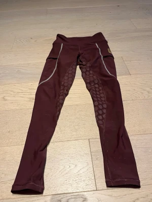 Maya Delores! Vinröda ridleggings med mönster - Säljer ett par vinröda ridleggings med hög midja och silikonmönster längs insidan av benen för extra grepp. De har fickor på sidorna och reflekterande detaljer. Perfekta för dig som gillar att rida och vill ha både stil och funktion.