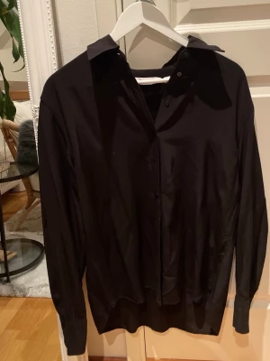 Svart skjorta med knappar hela vägen i strl 38 - Klassisk svart skjorta från Kappahl i storlek 38. Skjortan har lång ärm, knappar framtill och en stilren krage. Perfekt för dig som gillar en enkel och clean look. Material : Viskos 
