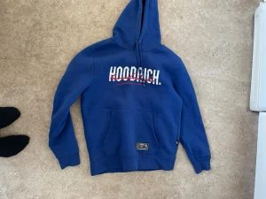 Blå hoodie från Hoodrich - Snygg blå hoodie från Hoodrich med stor logga på bröstet och ryggen. Klassisk känguruficka framtill och justerbar huva med dragsko. Tillverkad i mjukt material som känns skönt mot huden. Detaljer i vitt och rött på trycket samt patch nertill.