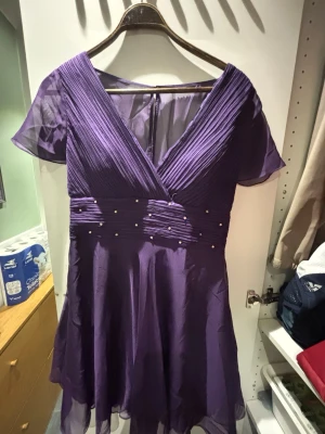 Lila klänning med pärlor - Snygg lila kortklänning från Ever Pretty med omlottdesign och v-ringning. Klänningen har korta, transparenta ärmar och ett plisserat liv med dekorativa pärlor i midjan. Tillverkad i ett lätt, flytande material som ger en elegant look. Storlek eu 44 tror jag och pris kan diskuteras 🤍