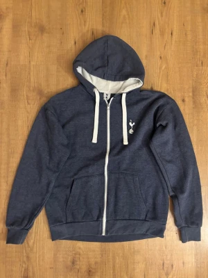 Blå zip-hoodie Tottenham Hotspur officiel merch - Mörkblå hoodie med dragkedja från Tottenham Hotspur. Tröjan har vit logga på bröstet, vita snören i huvan och vita detaljer vid dragkedjan. Mjuk insida och två fickor framtill. Perfekt för dig som gillar sportig stil och vill visa ditt lag.