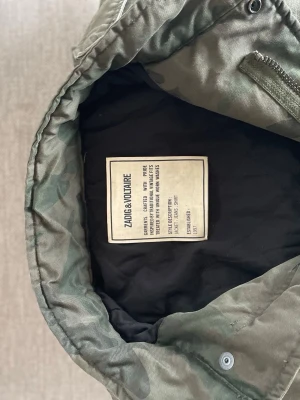 Field jacka från Zadig & Voltaire - Säljer en grön camo field jacka från Zadig & Voltaire med klassisk militärstil. Jackan har knappar på ärmslut, axelklaffar och flera fickor. Materialet är slitstark bomull och passformen är rak och avslappnad. Perfekt för lager-på-lager och streetwear. J