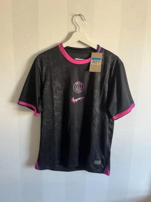 Svart PSG x Nike t-shirt med rosa detaljer - Säljer en svart PSG x Nike t-shirt med subtilt blommönster och rosa detaljer vid krage och ärmslut. Tröjan har korta ärmar och är tillverkad i ett lätt, syntetiskt material som passar perfekt till träning eller fotboll.