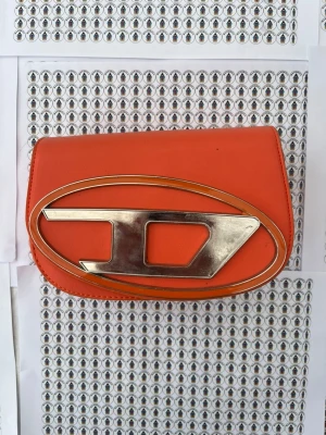 Orange kuvertväska med metallogo - Snygg orange kuvertväska i slätt material med stor silverfärgad metallogo på framsidan. Väskan har magnetlås och ett huvudfack. Perfekt för dig som vill sticka ut med en färgstark accessoar.