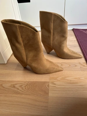 Beiga cowboy boots i mocka - Snygga cowboy boots i beige mocka med spetsig tå och hög klack. Bootsens höga skaft och stilrena design gör dem till ett statement-piece. Perfekta för dig som vill sticka ut med en trendig westernstil.