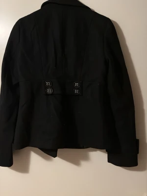 Svart rock från Lindex med knappar - Stilren svart kavaj från Lindex med dubbelknäppning framtill och dekorativa knappar i ryggen. Klassisk krage och breda slag, samt uppvikta ärmslut. Perfekt för dig som vill ha en tidlös och snygg look till hösten eller vintern.