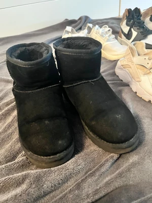 Uggs - Ett par svarta boots med kort skaft och rund tå. Utsidan är i mjuk mocka och insidan är fodrad med fluffigt material för extra värme. Klassisk och enkel design utan synligt märke. Perfekta för kalla dagar och riktigt sköna att ha på sig.