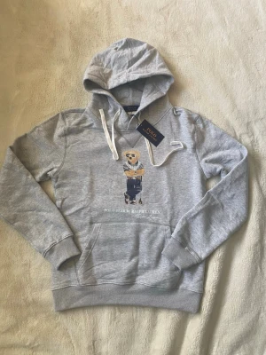 Grå hoodie Polo Ralph Lauren Björn - Säljer en grå hoodie från Polo Ralph Lauren med den ikoniska björnen på bröstet. Hoodien har vit dragsko i huvan, magficka och ribbade muddar. Tillverkad i mjukt bomullsmaterial och perfekt för en avslappnad streetwear-look.