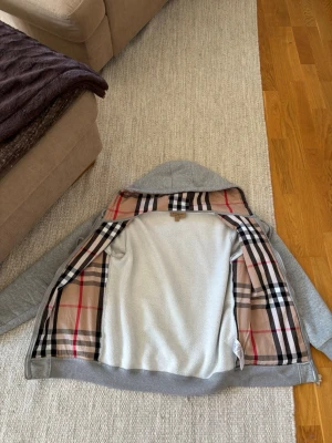 Grå hoodie från Burberry med dragkedja - Säljer en grå hoodie från Burberry med klassiskt rutigt foder i beige, svart och rött. Hoodien har dragkedja framtill, två fickor och justerbar huva med snören. Tillverkad i mjukt bomullsmaterial och har ribbade muddar vid ärmslut och nederkant. Lite mindre än vanlig M i storleken. Finns påse från burberry!