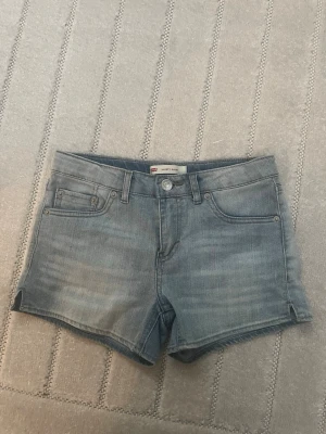 Blå jeansshorts från Levi's  - Säljer ett par klassiska blå jeansshorts från Levi's med fem fickor, bälteshällor och knappgylf. Shortsen har en ljus tvätt och små slitsar i sidorna. Skulle säga att de är i storlek xxxs/xxs