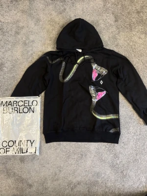 Svart hoodie Marcelo Burlon ormtryck - Svart hoodie från Marcelo Burlon County of Milan med unikt ormtryck i grönt, rosa och vitt på framsidan. Klassisk huva med dragsko och ribbade muddar. Materialet är mjuk bomull som känns skön mot huden. Perfekt för dig som vill sticka ut med en streetwear-look.
