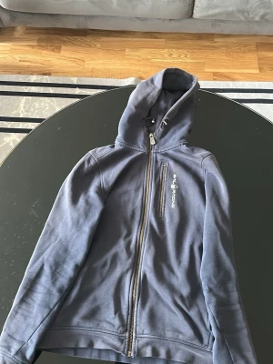 Blå zip-hoodie från Sail Racing - Snygg blå hoodie från Sail Racing med dragkedja framtill och en bröstficka med logga. Hoodien har justerbar huva och är tillverkad i mjukt bomullsmaterial. Perfekt för en avslappnad och sportig stil.