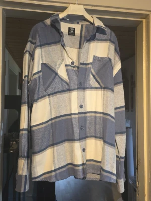 Blåvit oversize skjorta från FSBN - Oversize skjorta blått och vitt rutmönster från FSBN. Skjortan har två bröstfickor med lock, knäppning framtill och klassisk krage. Tillverkad i ett mjukt, vävt material som ger en avslappnad look. Perfekt att bära öppen över en t-shirt.
