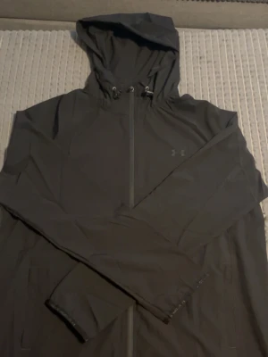 Svart vindjacka från Under Armour - Snygg svart vindjacka från Under Armour med huva och dragkedja framtill. Jackan har långa ärmar, justerbar huva med snörning och diskret logga på bröstet. Perfekt för blåsiga dagar och sportiga outfits.