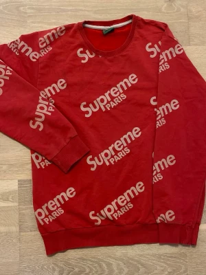 Röd Supreme Paris sweatshirt - Röd sweatshirt från Supreme Paris med vitt logotryck över hela tröjan. Rund halsringning, ribbade muddar vid ärmslut och nederkant. Tillverkad i mjukt bomullsmaterial för en avslappnad och trendig look.