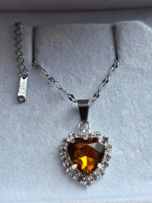 Halsband med hjärthänge och kristaller - Snyggt halsband i silverfärgad rostfritt stål med ett hjärtformat hänge i bärnstensfärgad kristall. Hänget är omgivet av gnistrande klara stenar som ger en lyxig känsla. Kedjan har en tvinnad design och justerbar längd. Perfekt för dig som gillar statement-smycken.