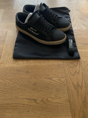 Svarta Saint Laurent sneakers - Svarta sneakers från Saint Laurent med vit text på sidan och beige sula. Klassisk låg modell med snörning och ovandel i läder. Kommer med svart dustbag och extra skosnören. Perfekta för en stilren och trendig look.