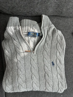 Grå kabelstickad tröja Polo Ralph Lauren - Snygg grå kabelstickad tröja från Polo Ralph Lauren med hög krage och dragkedja. Tröjan har långa ärmar och ribbade muddar vid ärmslut och nederkant. Perfekt för dig som gillar klassisk och stilren design. Storlek L/M. Bra skick. Storleks lappen är lite loss som synns på bilden