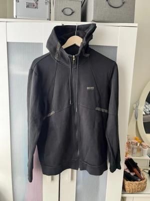 Hugo Boss Hoodie / kofta / luvtröja med zip - En mycket fin Hugo Boss huvtröja med dragkedja/ kofta med zip Svart färg med guldiga detaljer  Storlek M på lappen I nyskick Skicka meddelande vid några funderingar Kolla gärna även in mina andra annonser