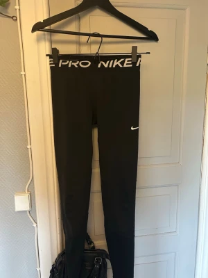 Svarta Nike Pro tights - Säljer ett par svarta Nike Pro tights med vit logga på benet och bred resår i midjan med texten 'NIKE PRO'. Tightsen är i stretchigt material som passar perfekt till träning och sportaktiviteter. Snygg och enkel design som är lätt att matcha. De är i JR L men skulle säga att det är som xs!🥰