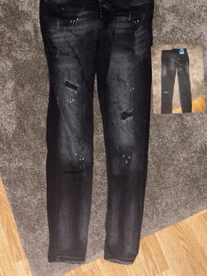 Dsquared byxor  - Säljer ett par svarta jeans med slitningar och coola färgstänk på framsidan. Jeansen har rak passform och detaljerade revor på båda benen för en trendig look. Perfekta för dig som gillar streetstyle och vill sticka ut lite extra.
