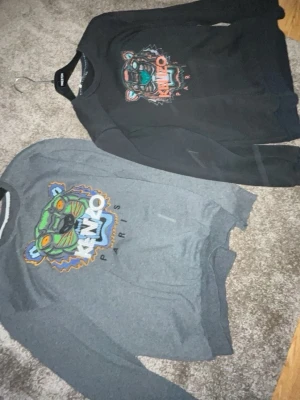 Kenzo sweatshirts med tigertryck - Varsin pris för varsin tröja Två snygga sweatshirts från Kenzo med ikoniskt tigertryck på bröstet. En är svart och den andra är grå, båda med färgglada detaljer i trycket och texten 'KENZO PARIS'. Klassisk rund hals och mjukt material, perfekta för en trendig streetwear-look.