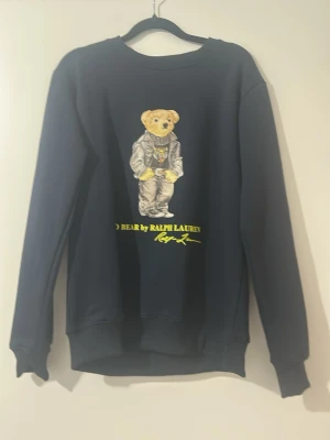 Mörkblå sweatshirt Polo Ralph Lauren - Säljer en mörkblå sweatshirt från Polo Ralph Lauren med den ikoniska björnen på bröstet. Tröjan har rund halsringning, ribbade muddar och är gjord i mjukt bomullsmaterial. Perfekt för dig som vill ha en klassisk och stilren look med en twist.