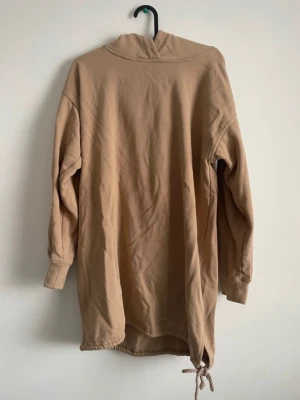 Beige oversized hoodie med dragsko - Säljer en beige oversized hoodie med huva och dragsko längst ner. Hoodien har långa ärmar och ribbade muddar vid ärmsluten. Perfekt för en chill och avslappnad stil. Materialet känns mjukt och skönt, troligen bomull eller bomullsblandning.