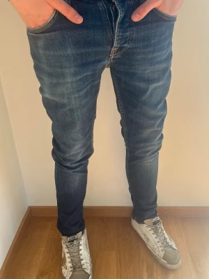 Mörkblå slim fit jeans från Nudie - Säljer ett par mörkblå slim fit jeans från Nudie Jeans med snygga slitningar och kontrastsömmar. Jeansen har klassisk femficksmodell, orange broderi på bakfickorna och är tillverkade i stretchig denim för skön passform. Storleken står nedan. Perfekta nu till våren. Hör av er vid frågor! 