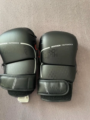 Svarta boxningshandskar Outshock - Ett par svarta mma handskar från Decathlon, modell Outshock. Handskarna har kardborreband runt handleden för enkel justering och sitter skönt på handen. Tillverkade i slitstarkt syntetmaterial med diskreta vita detaljer och logga på ovansidan.