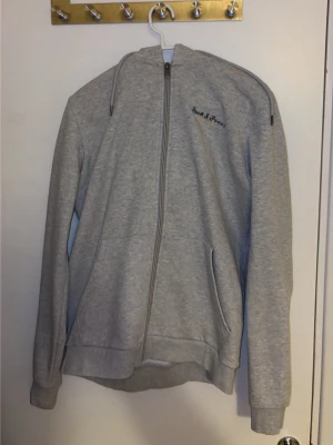 Grå zip-hoodie från Jack & Jones - Grå hoodie med dragkedja från Jack & Jones. Klassisk modell med huva och dragsko, två fickor framtill och broderad logga på bröstet. Mjuk bomullsblandning som är skön att ha på sig. Perfekt för en avslappnad stil. S men passar M