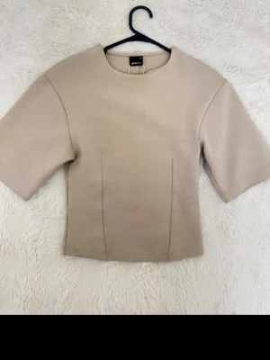Beige boxy topp från Gina - Stilren beige topp från Monki med boxig passform och halvlånga, vida ärmar. Tillverkad i ett tjockare, mjukt material som ger en clean look. Rund halsringning och diskreta sömdetaljer framtill för extra edge.
