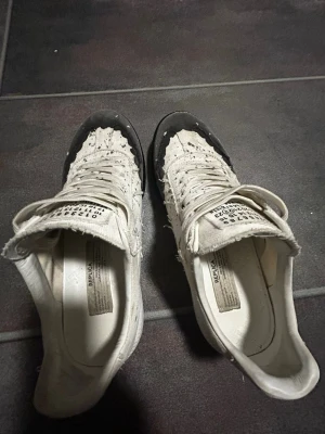 Maison Margielas - Unika vita sneakers från Maison Margiela med svart tåparti och coola färgstänk över hela skon. Låg modell med slitna detaljer och klassisk snörning. Ovandelen är i tyg och läder, och tungan har märkets signatur-etikett.