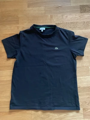 Svart t-shirt från Lacoste - Klassisk svart t-shirt från Lacoste med den ikoniska gröna krokodilloggan broderad på bröstet. T-shirten har rund halsringning och korta ärmar. Tillverkad i mjuk bomull för en skön känsla och enkel stil. Perfekt till våren och sommaren.