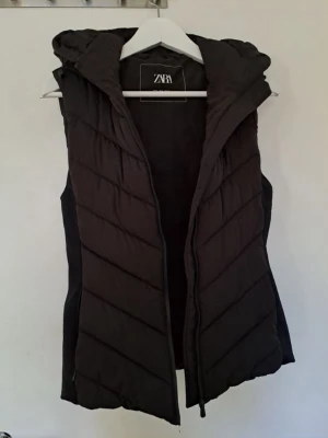 Svart dunväst med huva från Zara - Snygg svart dunväst från Zara med dragkedja framtill och stor, vadderad huva. Har bara använt ett få par gånger så den är i väldigt bra skick. Västen är perfekt för våren men är tyvärr för liten för mig och därför säljer jag den.